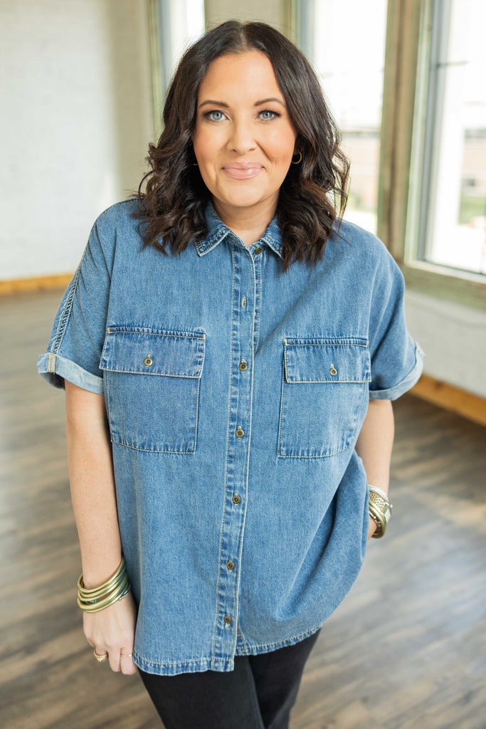 Plus-size woman wearing a medium-wash denim button up top with rolled cap sleeves and chest pockets.
