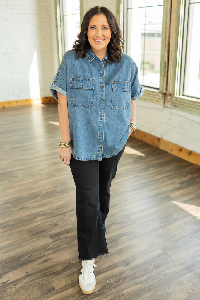 Plus-size woman wearing a medium-wash denim button up top with rolled cap sleeves and chest pockets.