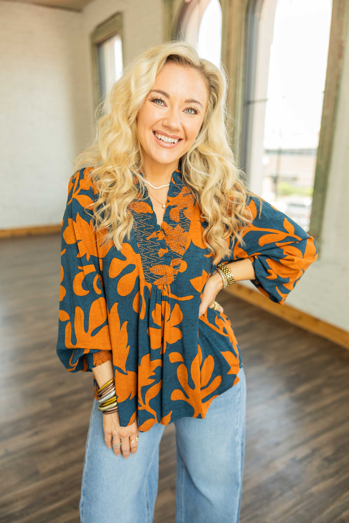 Blonde woman wearing navy and burnt orange printed long sleeve challis top with smocked neckline and balloon sleeves.