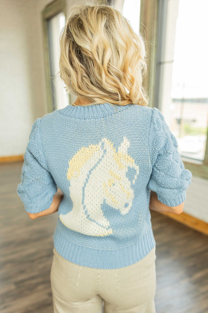 Woman wearing a vintage-inspired blue short sleeve knit cardigan with horseshoe details on pockets and a horse design on the back.