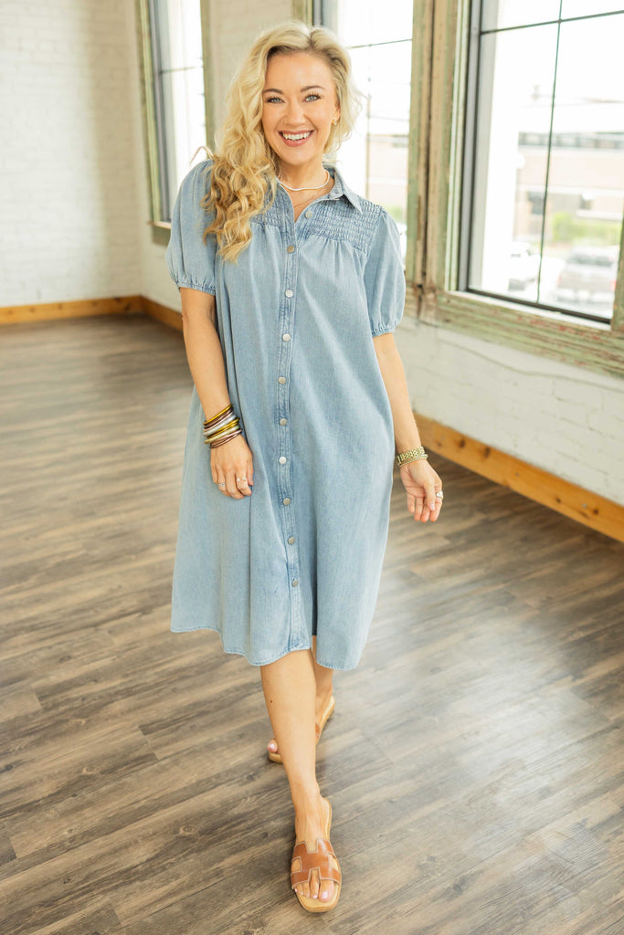 ALT text suggestion: Blonde woman wearing the Savanna Denim Midi Button Up Dress with short puff sleeves and front buttons, styled with tan sandals, standing in a bright room with wood floors.