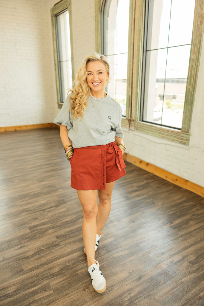 Woman wearing gray oversized embroidered smiley face t-shirt styled casually with a rust skort and sneakers.