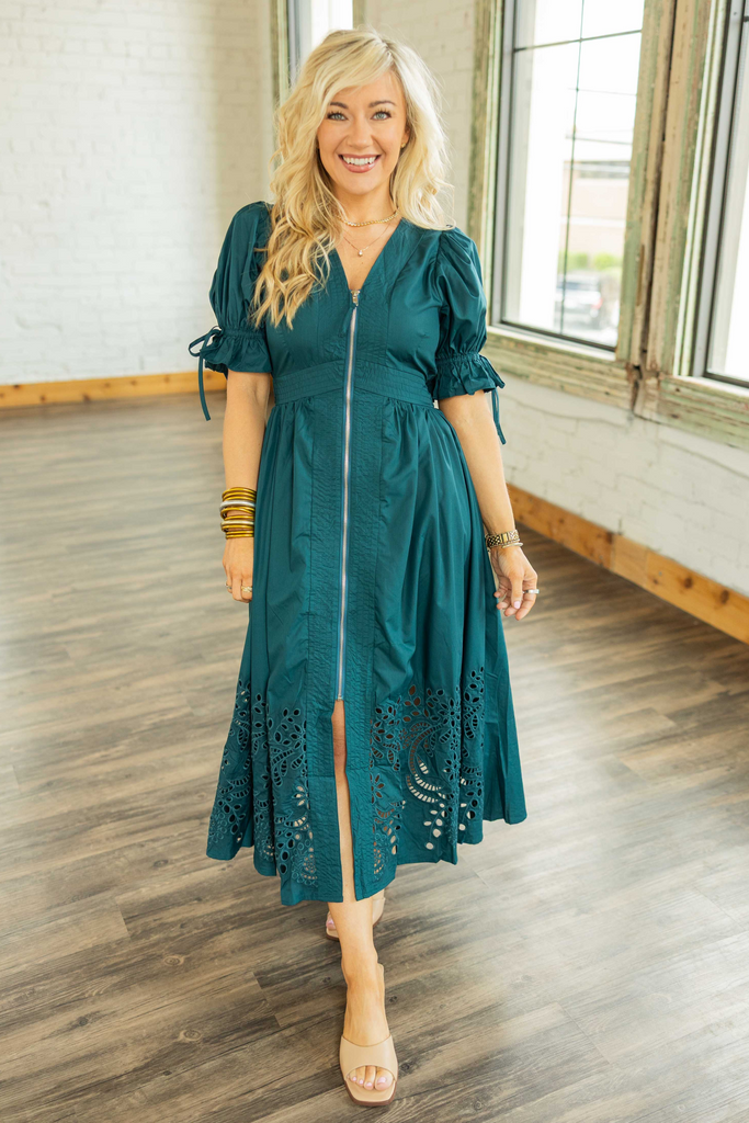 Blonde woman wearing a deep teal puff sleeve midi dress with zipper front and embroidered hem.