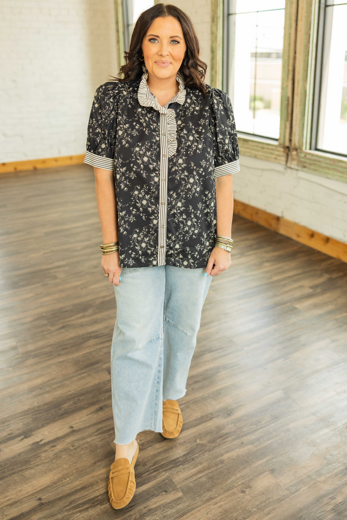 Woman wearing a black floral button up top with striped ruffle neckline and short puff sleeves, styled casually with jeans.