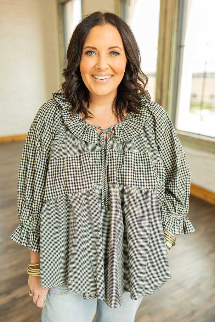 Woman wearing a plus-size black and ivory gingham top with ruffle sleeves, tie-front neckline, and high-low hem.