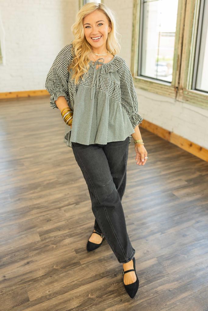 Woman wearing a black and ivory gingham top with ruffle 3/4 sleeves, front tie detail, and high-low hem.