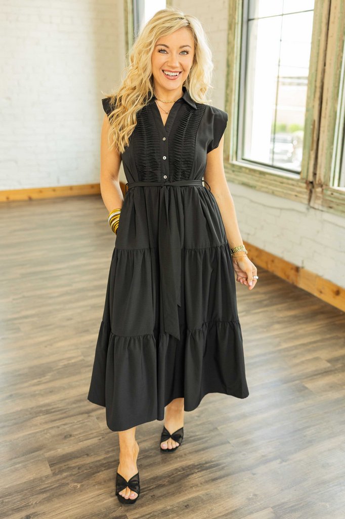 Blonde woman wearing a black sleeveless midi dress with pleated front detail, waist tie, and tiered skirt.