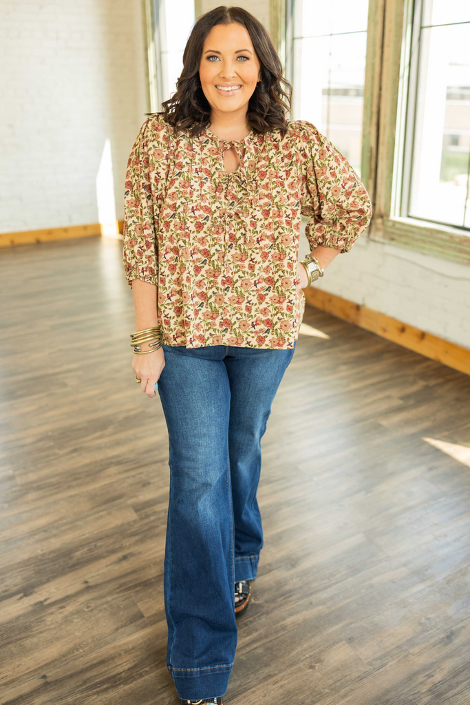 plus size curvy woman wearing a cream corduroy floral pintuck blouse with a tie neck keyhole and 3/4 sleeves, featuring red rose and green vine print with small blue birds, paired with jeans indoors