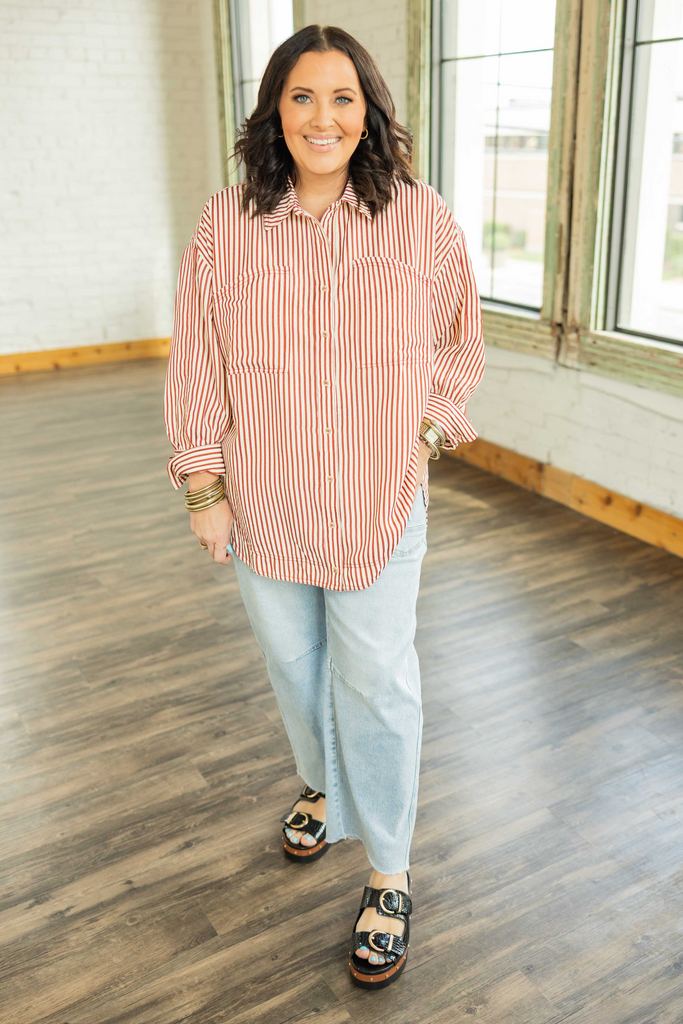 CURVY Woman wearing a striped shirt and light jeans standing in a room with wooden flooring and large windows.