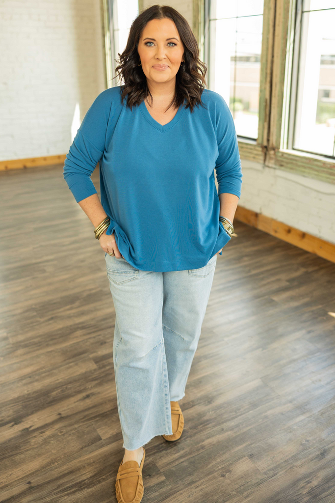 Woman wearing a blue sweater and light jeans standing in a room with wooden flooring and large windows.