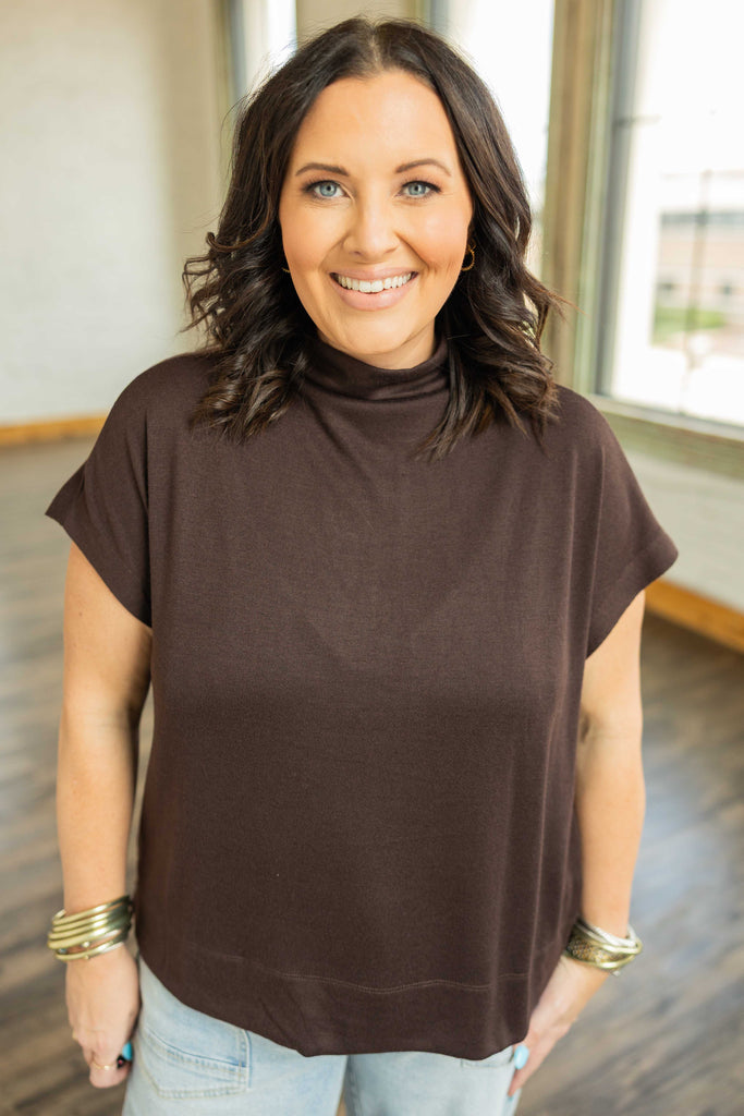 Woman wearing a chocolate brown mock neck pullover top with cap sleeves and light blue jeans