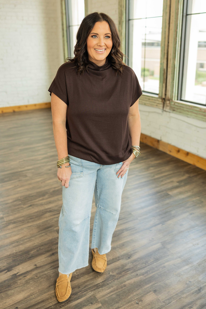 Woman wearing a chocolate brown mock neck pullover top with cap sleeves and light blue jeans