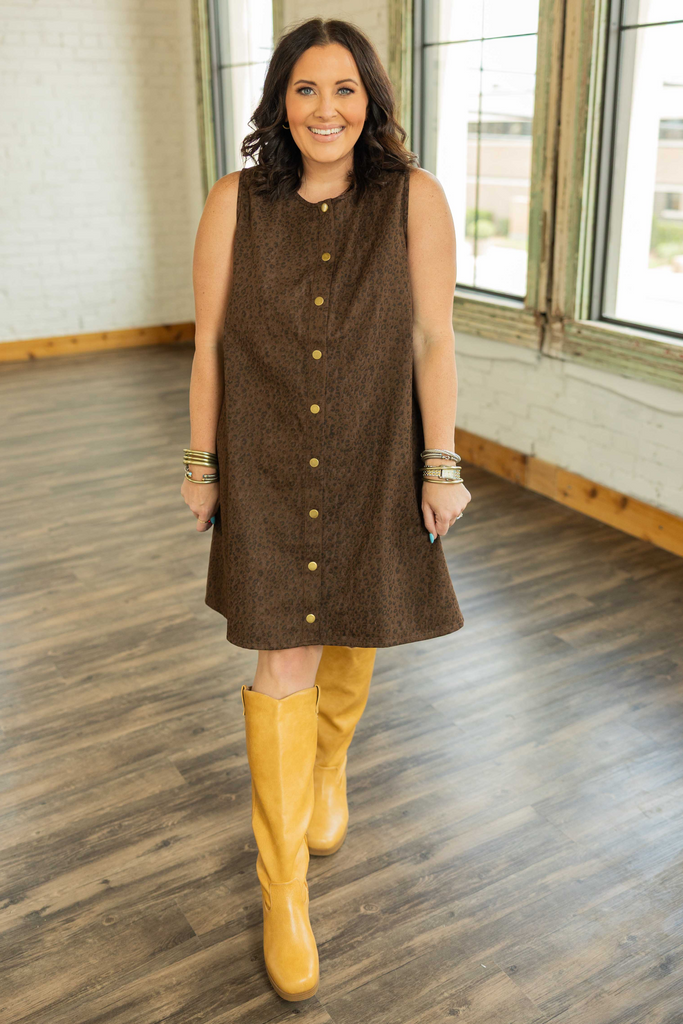 Woman in a chocolate brown sleeveless mini dress with subtle leopard print and gold snap buttons standing in a bright loft space.