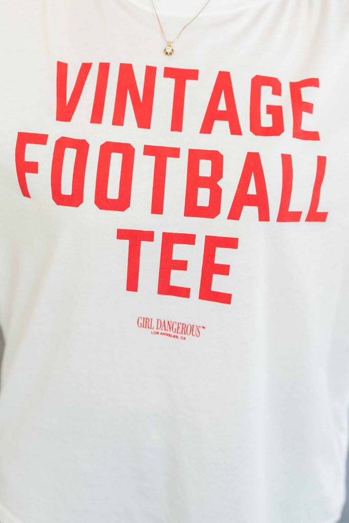 Blonde woman wearing a red oversized graphic tee with yellow “Game Day” lettering and vintage football-inspired cartoon graphics, paired with cuffed blue jeans and sandals, standing indoors with bright windows