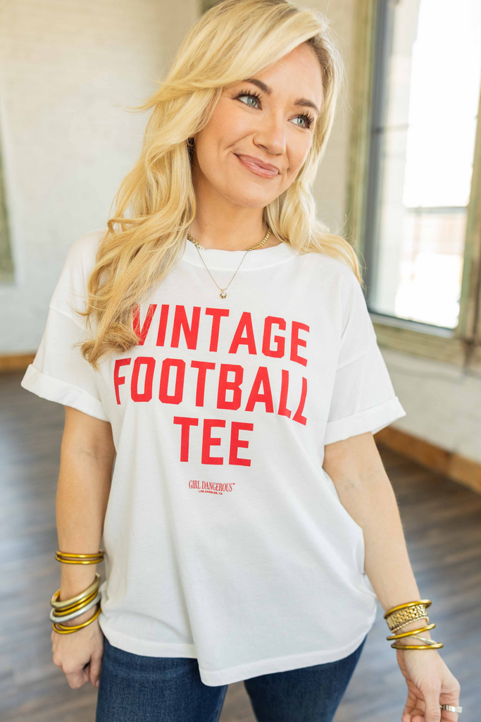 Blonde woman wearing a red oversized graphic tee with yellow “Game Day” lettering and vintage football-inspired cartoon graphics, paired with cuffed blue jeans and sandals, standing indoors with bright windows