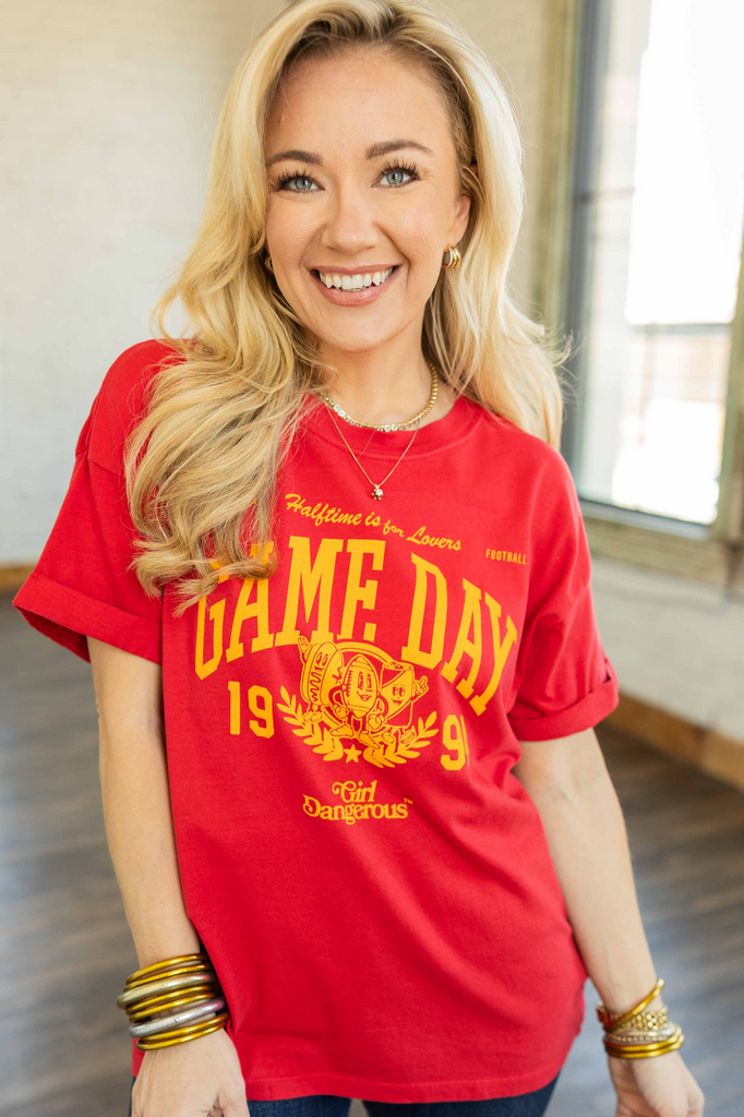 Blonde woman wearing a white oversized graphic tee with bold red “Vintage Football Tee” lettering, paired with cuffed blue jeans and layered gold bracelets, standing in a bright room with wood floors