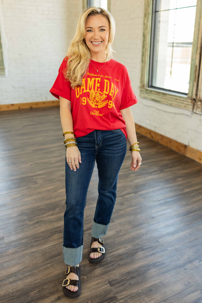 Blonde woman wearing a white oversized graphic tee with bold red “Vintage Football Tee” lettering, paired with cuffed blue jeans and layered gold bracelets, standing in a bright room with wood floors