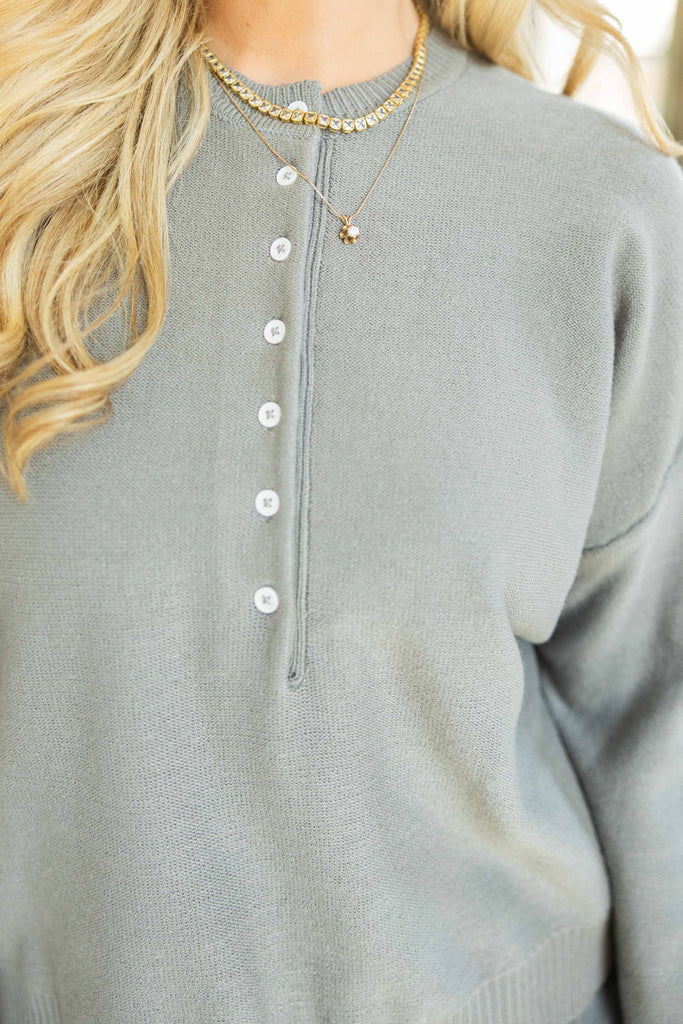 Close-up of a person wearing a light gray long-sleeve shirt with buttons, gold necklace, and blonde hair.