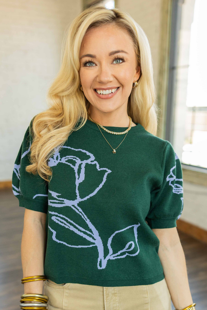 Blonde woman wearing a forest green cropped short sleeve sweater knit pullover with oversized lavender floral motif, styled with khaki wide leg pants indoors.