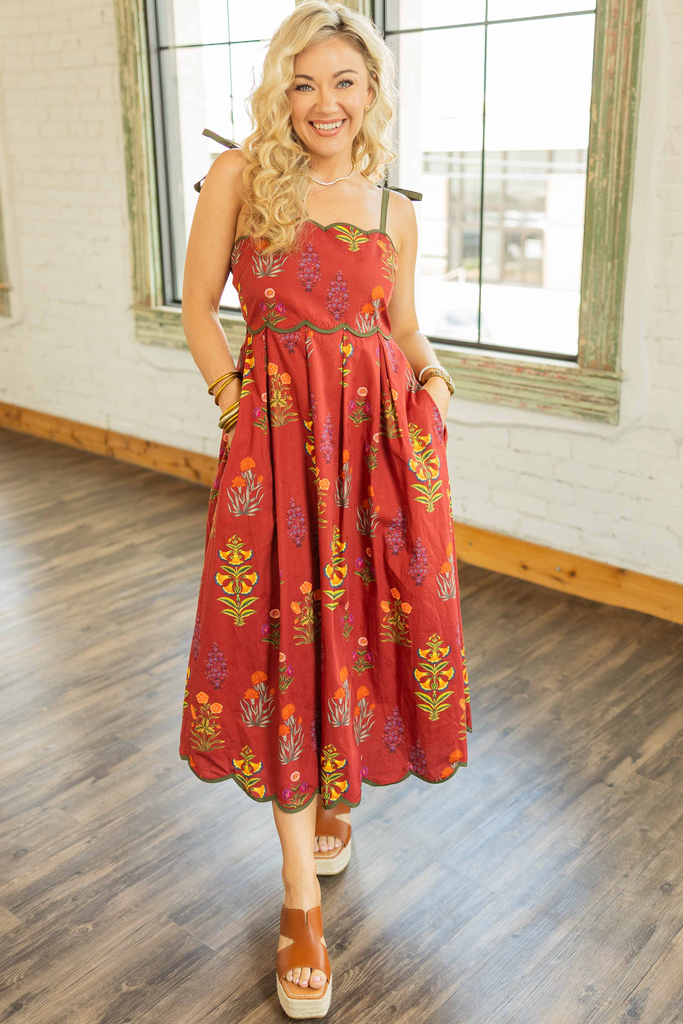 Woman wearing a brick red floral midi dress with scalloped neckline and tie straps, styled for fall in a rustic loft setting.