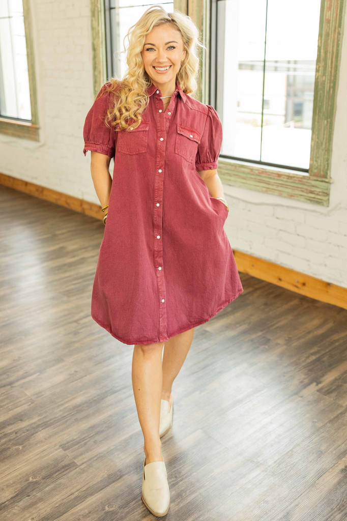 Woman wearing a cranberry denim mini dress with pearl snap buttons, puff sleeves, and front square pockets in a bright loft setting.