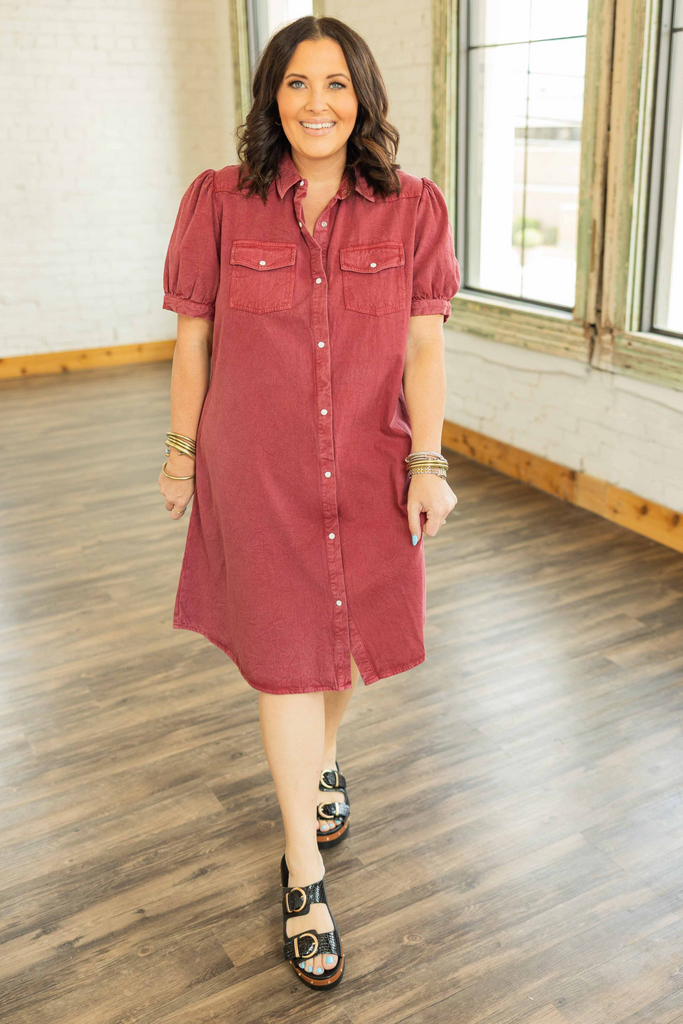 Woman wearing a cranberry denim mini dress with pearl snap buttons, puff sleeves, and front square pockets in a bright loft setting.