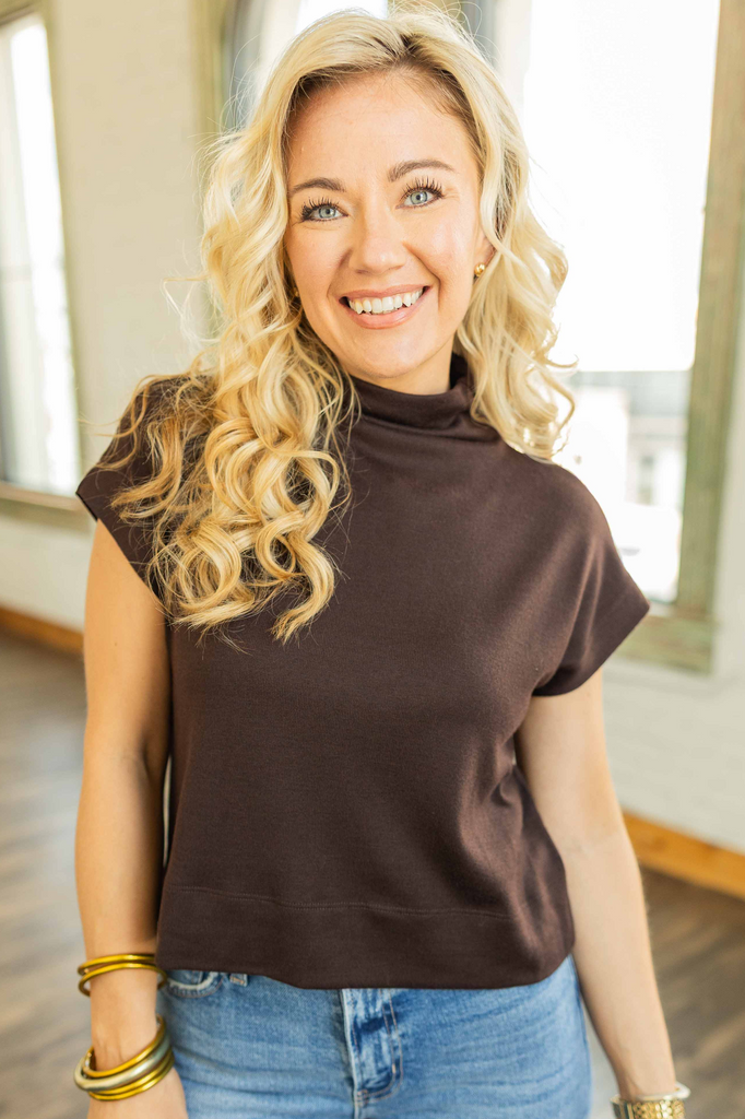 Woman wearing a chocolate brown mock neck pullover top with cap sleeves and light blue jeans