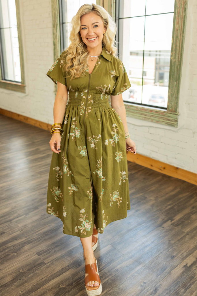 Woman wearing an olive green floral midi dress with short sleeves, smocked waist, and front zipper standing in a bright loft space.