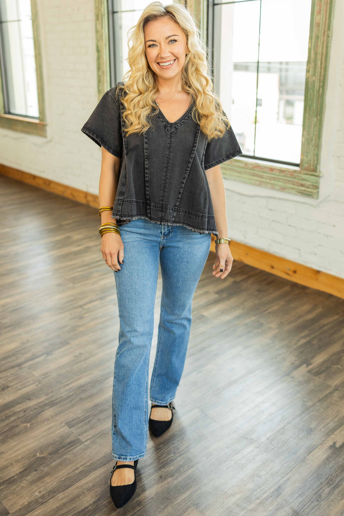 Woman wearing a washed black denim v-neck top with exposed seams and frayed hem styled with light wash jeans
