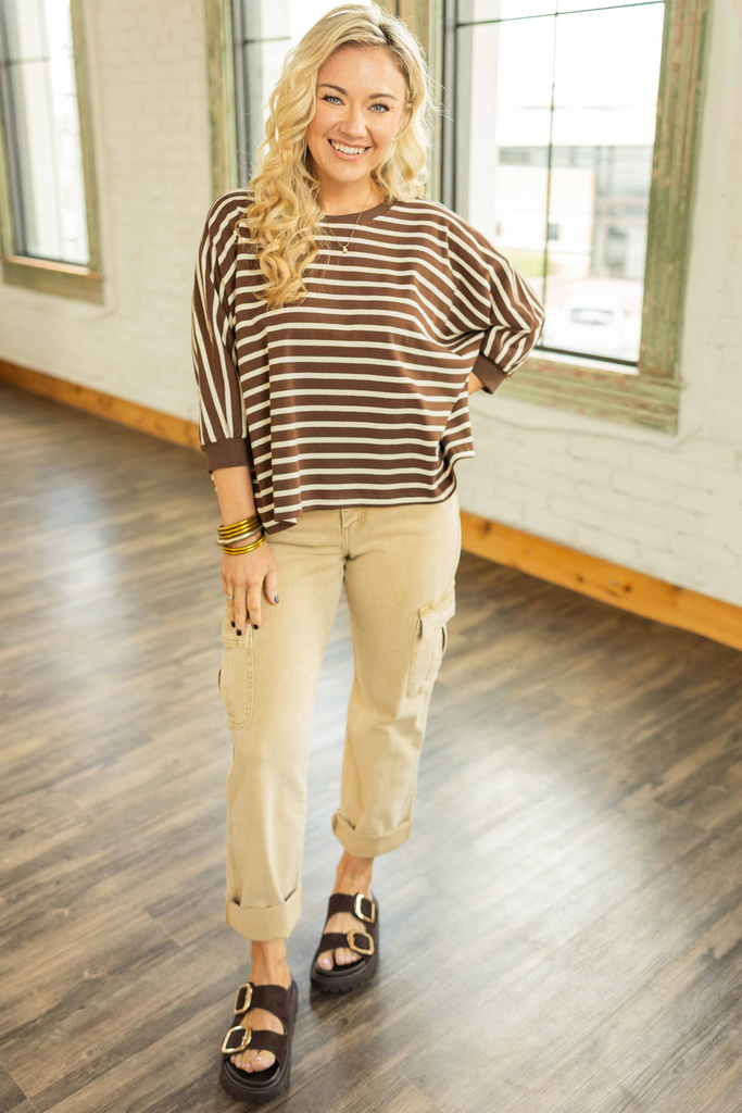 Woman wearing a chocolate brown and ivory striped oversized knit top with 3/4 sleeves styled with beige cargo jeans in a bright room.