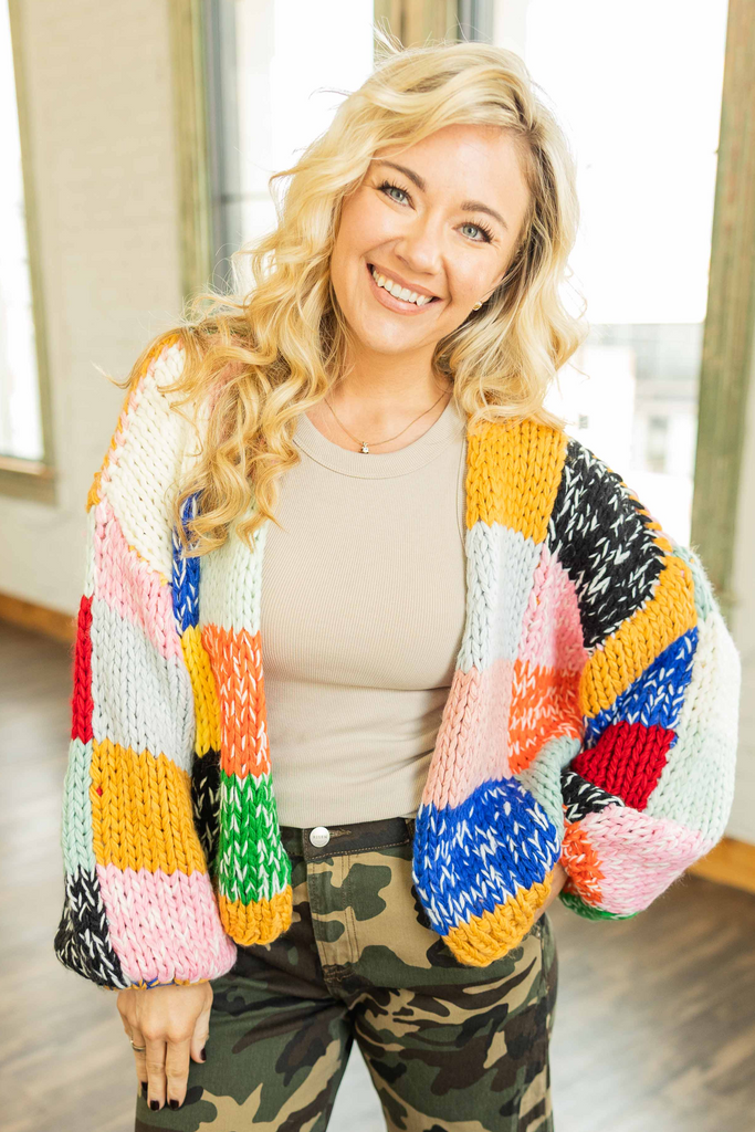 Woman wearing a chunky knit cardigan with bright color block panels of yellow, pink, red, green, blue, orange, and black standing in a bright loft space.