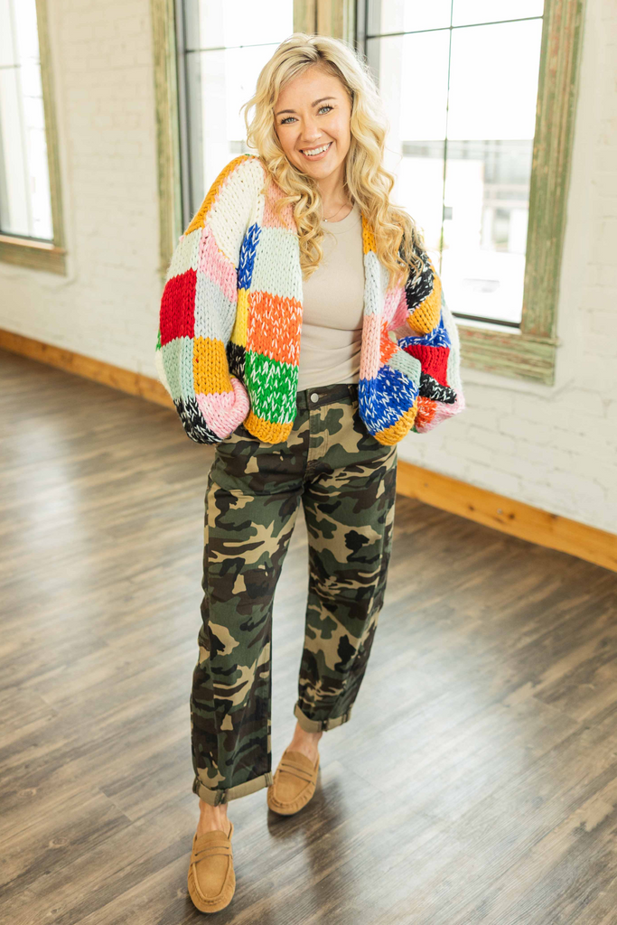 Woman wearing a chunky knit cardigan with bright color block panels of yellow, pink, red, green, blue, orange, and black standing in a bright loft space.