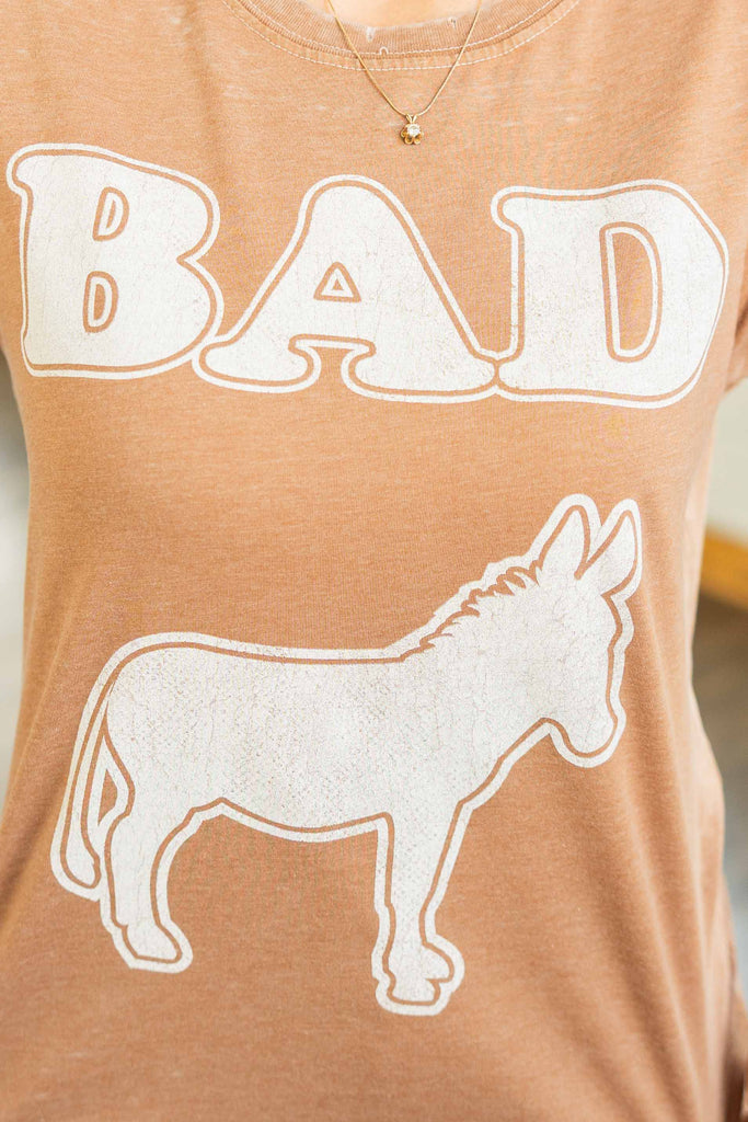 Woman wearing a mocha brown mineral washed graphic tee with “BAD” text and white donkey illustration