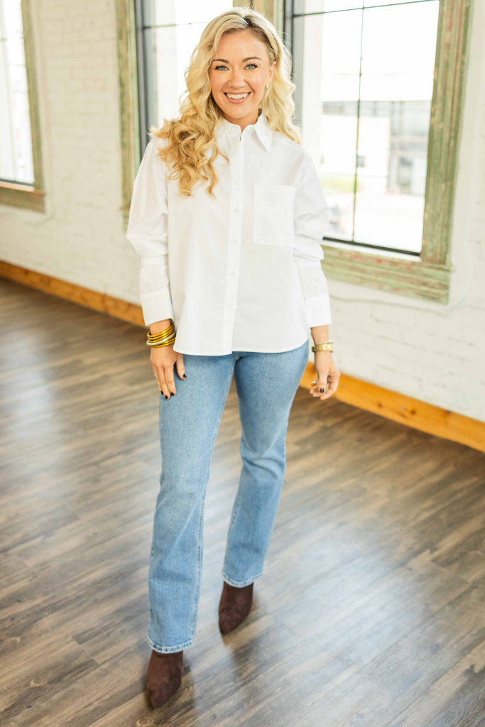 Woman wearing a crisp white button down top with bishop sleeves and a front pocket styled with light wash jeans in a bright loft space.