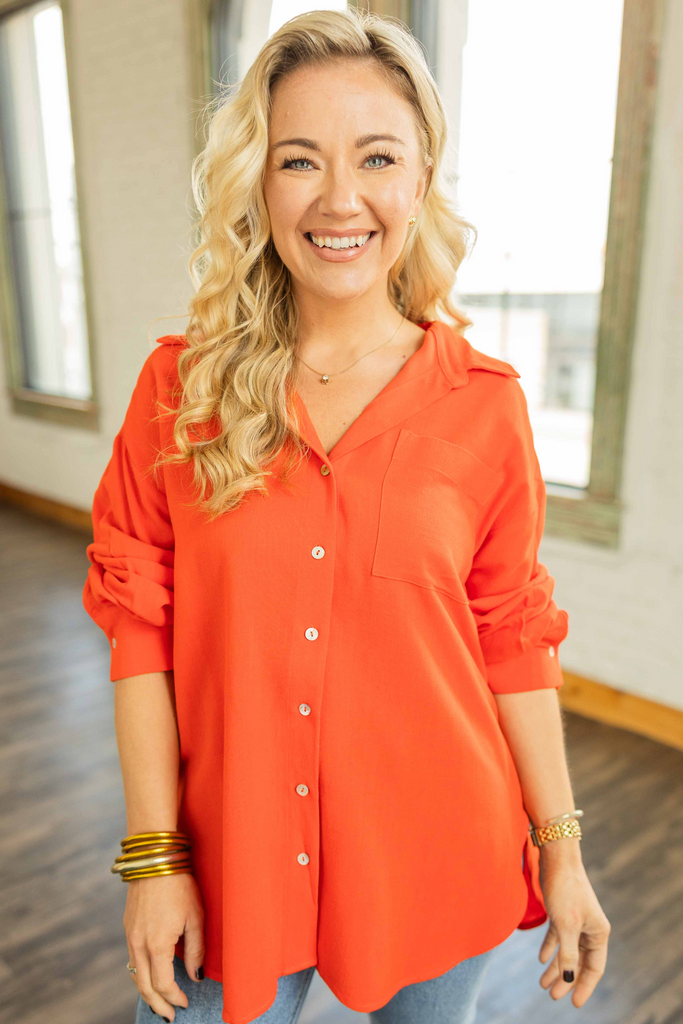 Woman wearing an oversized poppy red linen blend button up top with roll tab sleeves and collared neckline standing in a bright loft space.