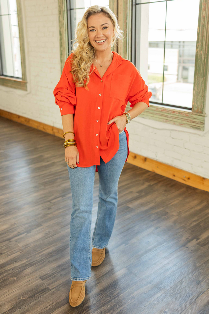 Woman wearing an oversized poppy red linen blend button up top with roll tab sleeves and collared neckline standing in a bright loft space.