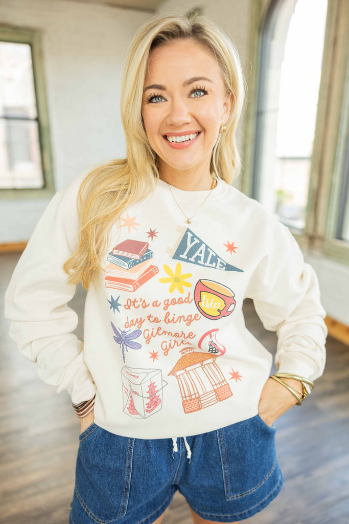 Woman wearing cream sweatshirt with colorful Gilmore Girls themed graphic including books, Luke’s coffee cup, and Stars Hollow gazebo, styled with denim shorts.