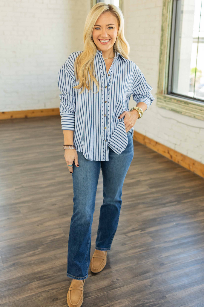Woman wearing oversized blue and white striped button-down top with long sleeves and pleated back hem detail, styled with jeans in a bright studio.