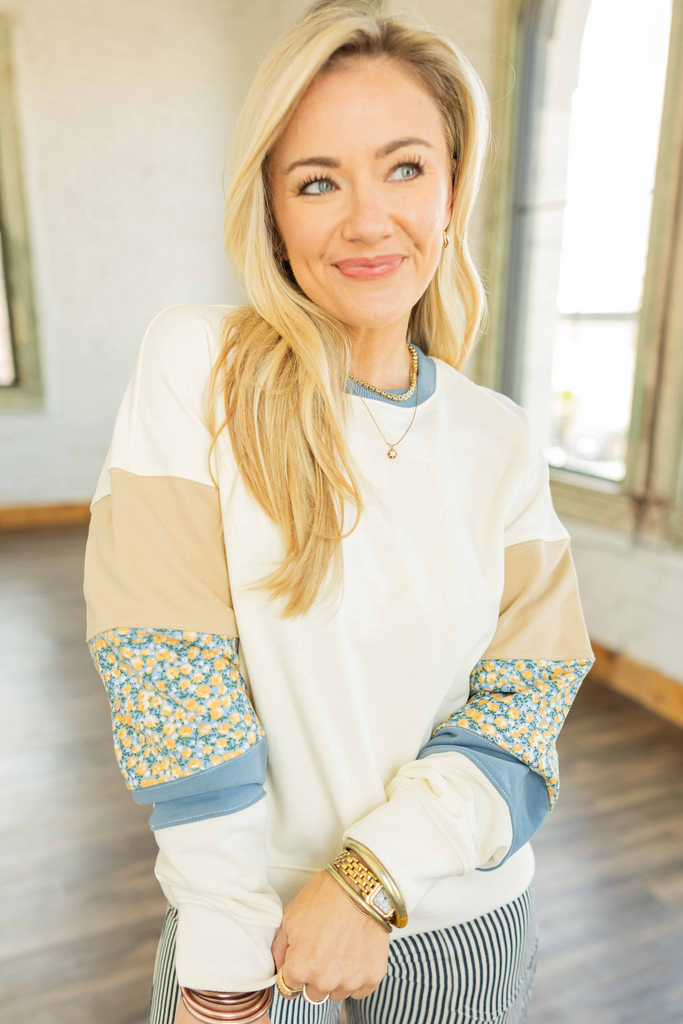 Woman wearing cream pullover top with multi-fabric patchwork sleeves in tan, blue, and floral prints, styled with striped pants in bright studio.