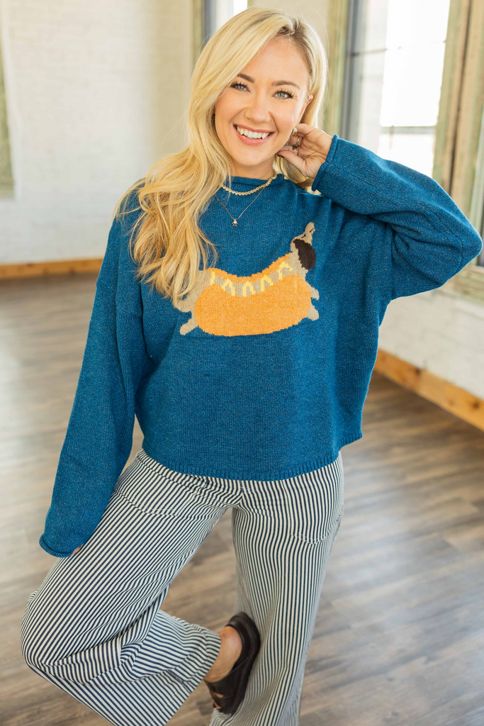 Woman wearing teal relaxed fit sweater with playful hot dog dachshund graphic, slight crop, and drop shoulders in bright studio.