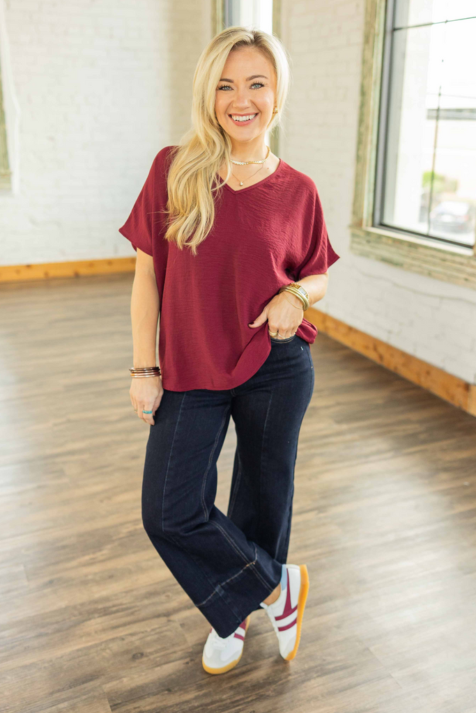 Woman wearing burgundy V neck short sleeve top with relaxed fit, paired with dark denim jeans and sneakers in bright studio.