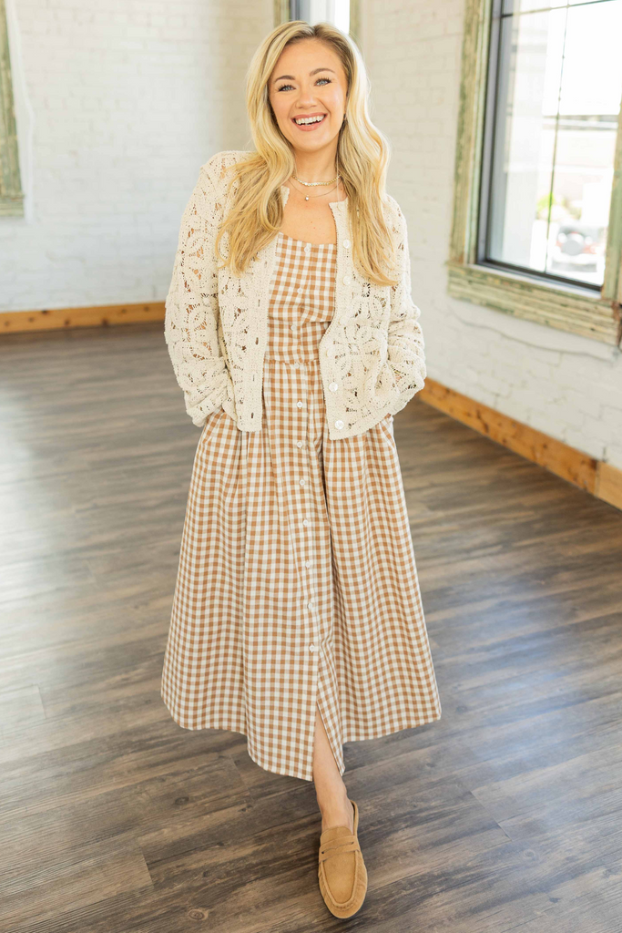 Woman wearing cream lace crochet cardigan over tan gingham dress, standing in bright studio with wood floors.