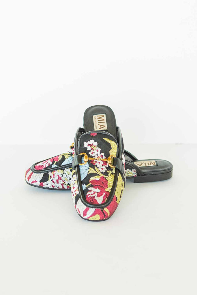 Pair of black slip-on mules with colorful floral embroidery and gold horse-bit detail on a white background.