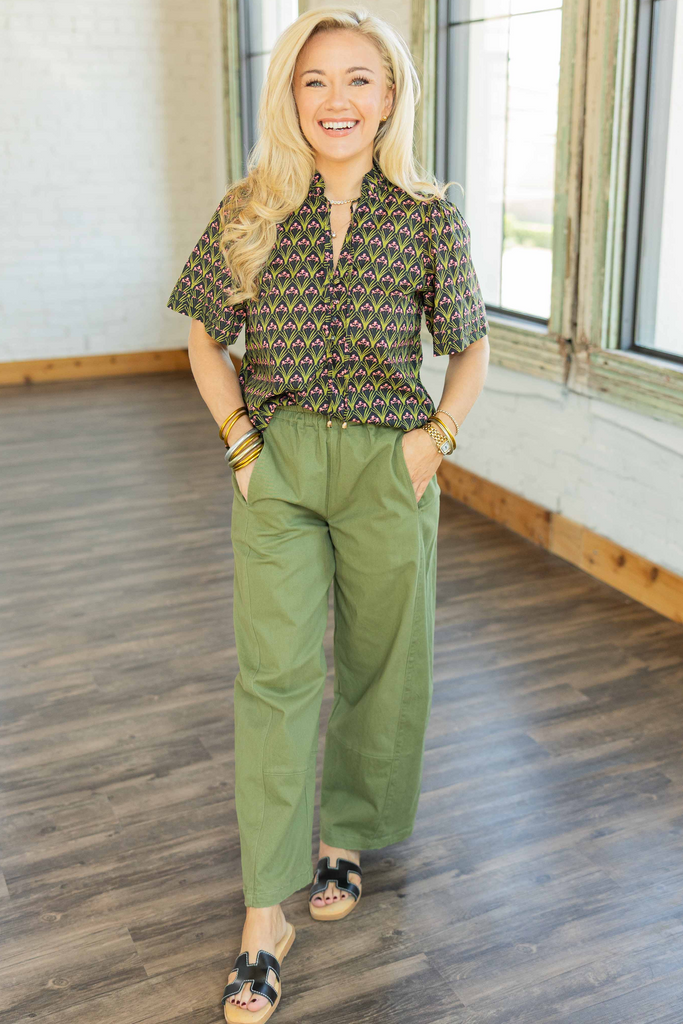 Woman wearing black top with green and pink geometric print, ruffled mock neck, and short sleeves styled with olive wide-leg pants.