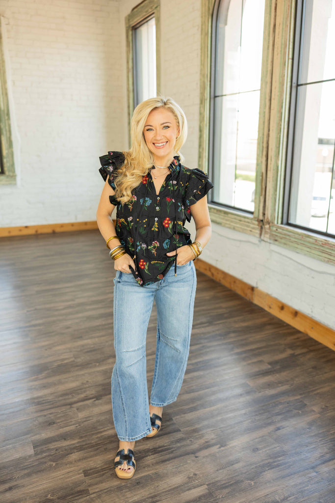 Woman wearing black flutter sleeve top with colorful floral embroidery and button-front neckline styled with light wash denim BARREL jeans.