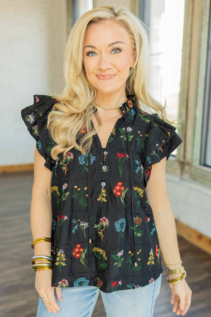 Woman wearing black flutter sleeve top with colorful floral embroidery and button-front neckline styled with light wash denim BARREL jeans.