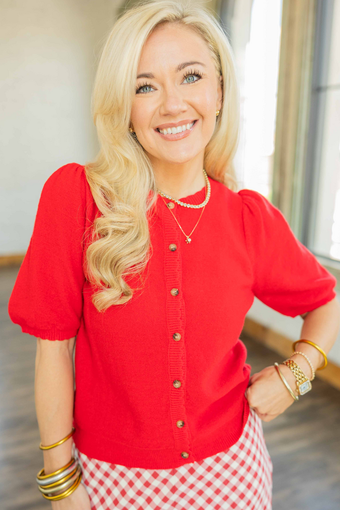 Woman wearing bright red button-up short sleeve cardigan top with scalloped trim and puff sleeves styled with red and white gingham skirt.