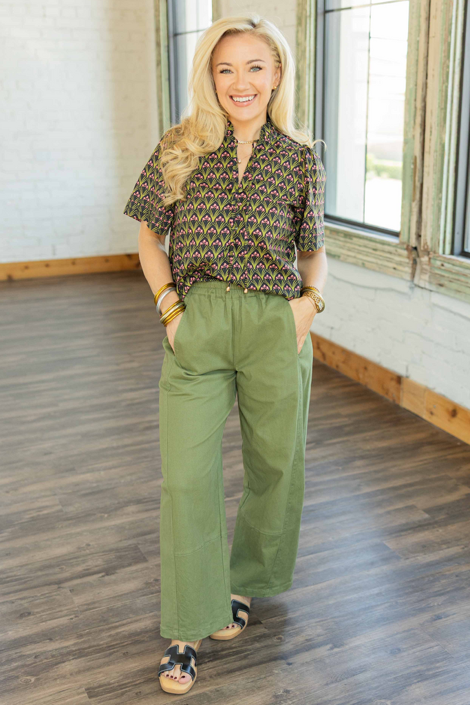 Woman wearing olive green cropped pants with an elastic waistband, styled with a printed blouse and sandals, standing indoors.