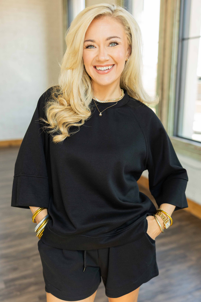 Woman wearing black 3/4 sleeve drop shoulder top with relaxed fit and crew neckline styled with black drawstring shorts.