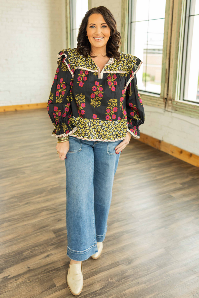 curvy plus Woman wearing black multi-print long sleeve top with ruffle accent shoulders, puff sleeves, and striped V-neck band styled with jeans